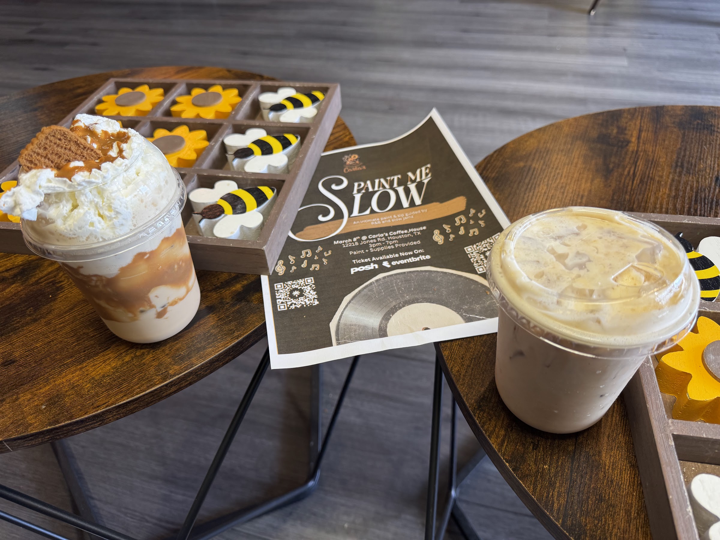 Iced coffee drinks, a Paint Me Slow event flyer, and bee-themed game on small tables.
