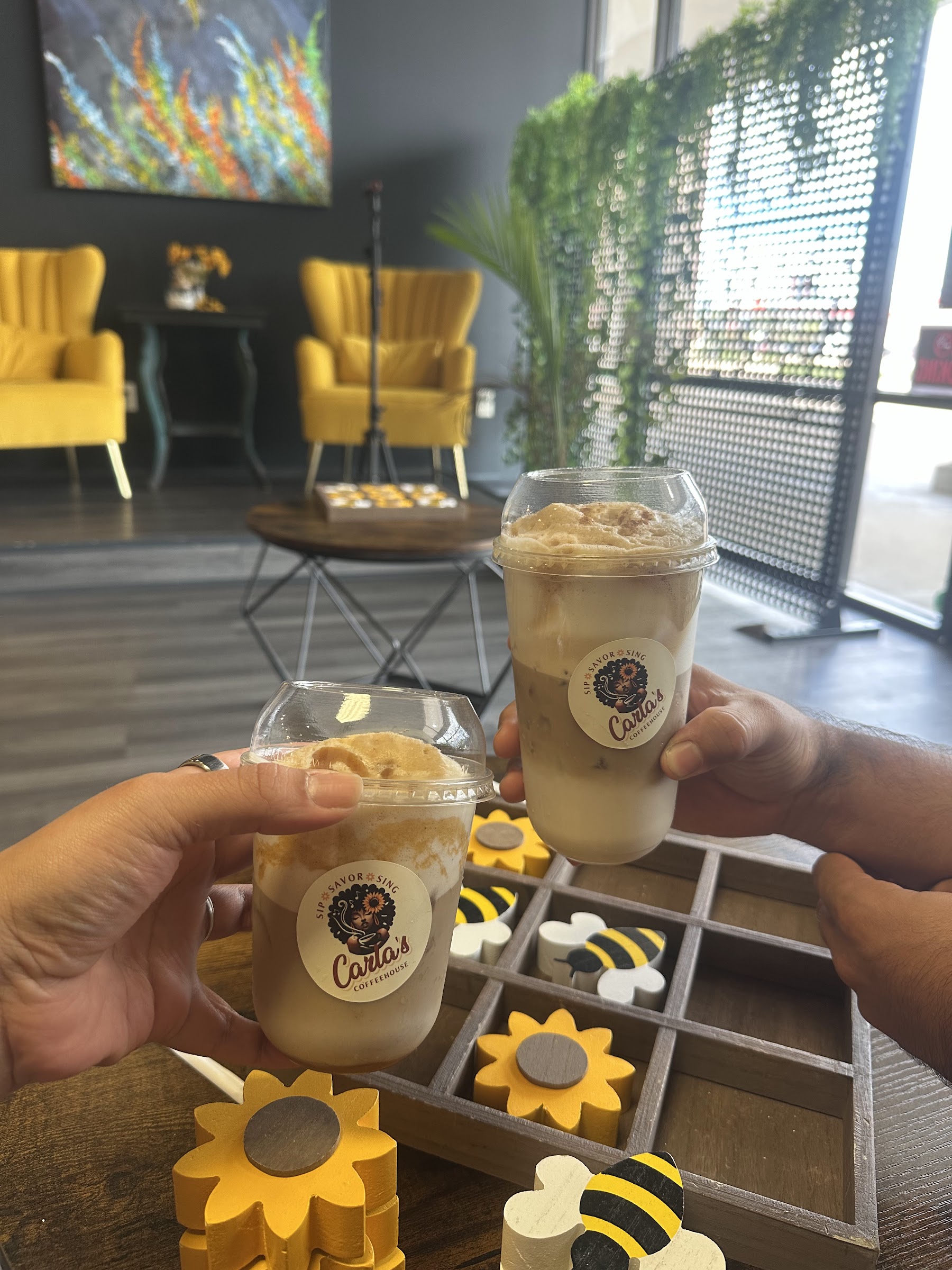 Two people toast with iced Carla's Coffeehouse drinks over a bee-themed game